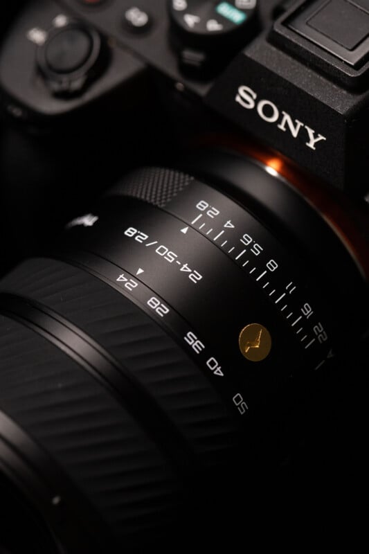 Close-up of a Sony camera showing detailed markings on the attached lens, including focal length numbers and focus indicators, with a metallic black finish and a gold accent visible.