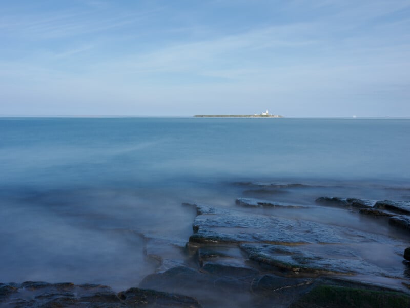 Rocky shoreline with gentle waves in the foreground, calm blue sea, and a distant island with a lighthouse under a clear sky.