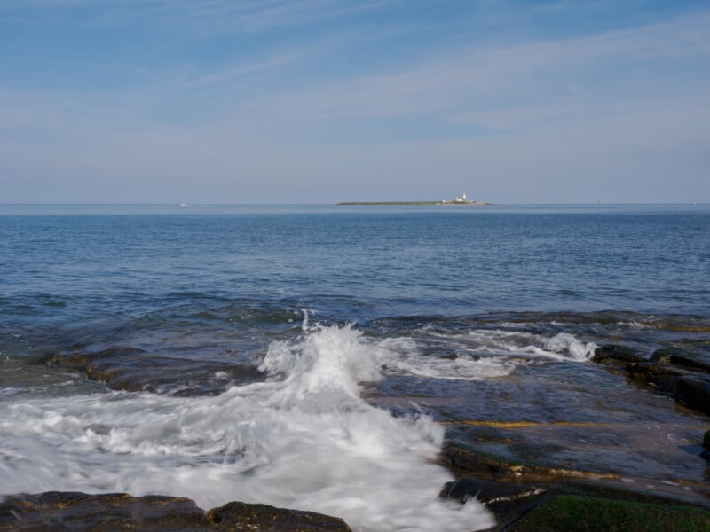 Waves crash against rocky shore in the foreground, with a calm blue sea and a distant island featuring a small lighthouse under a partly cloudy sky.