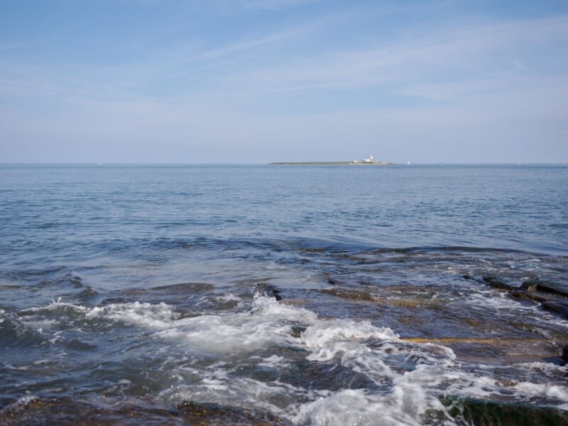 Waves gently crash on rocky shore in the foreground, with a calm blue sea and a distant small island featuring a white lighthouse under a partly cloudy sky.
