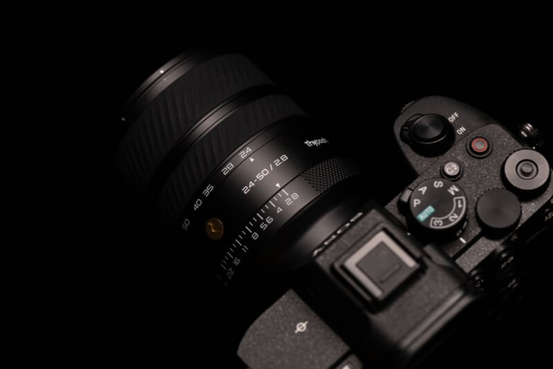 Close-up of a black digital camera with a zoom lens, showing detailed lens markings and part of the camera body with control dials, all set against a dark background.