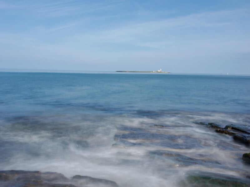Rocky shoreline with gentle waves in the foreground, calm blue sea, and a distant small island with a white lighthouse under a clear sky.