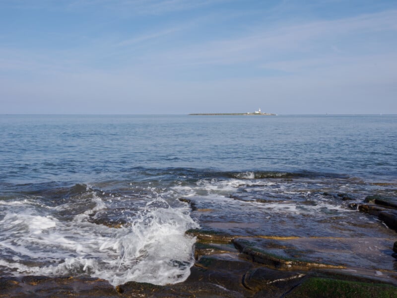 Waves splash against dark, rocky shore in the foreground. Calm blue sea stretches to the horizon with a small, distant island and lighthouse visible under a partly cloudy sky.