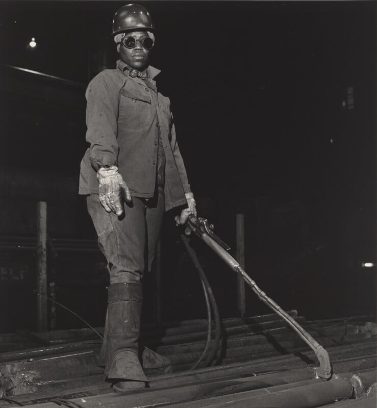 A factory worker wearing protective goggles, gloves, and a helmet stands on metal beams, holding industrial equipment in a dimly lit environment.