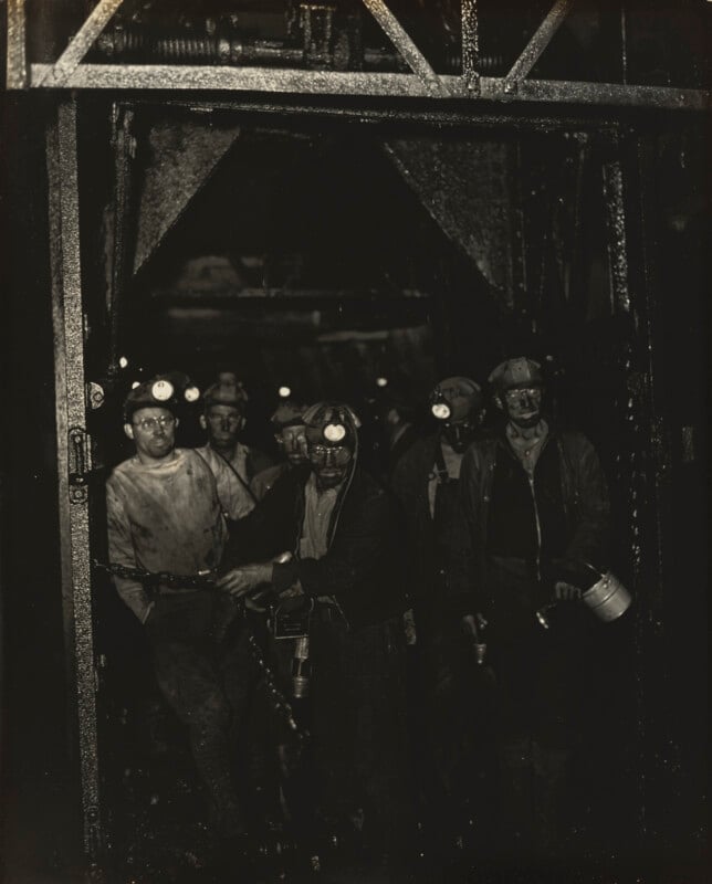 A group of coal miners with headlamps stand together in a dark, industrial setting, their faces and clothes covered in dirt, suggesting they have just finished a shift underground.