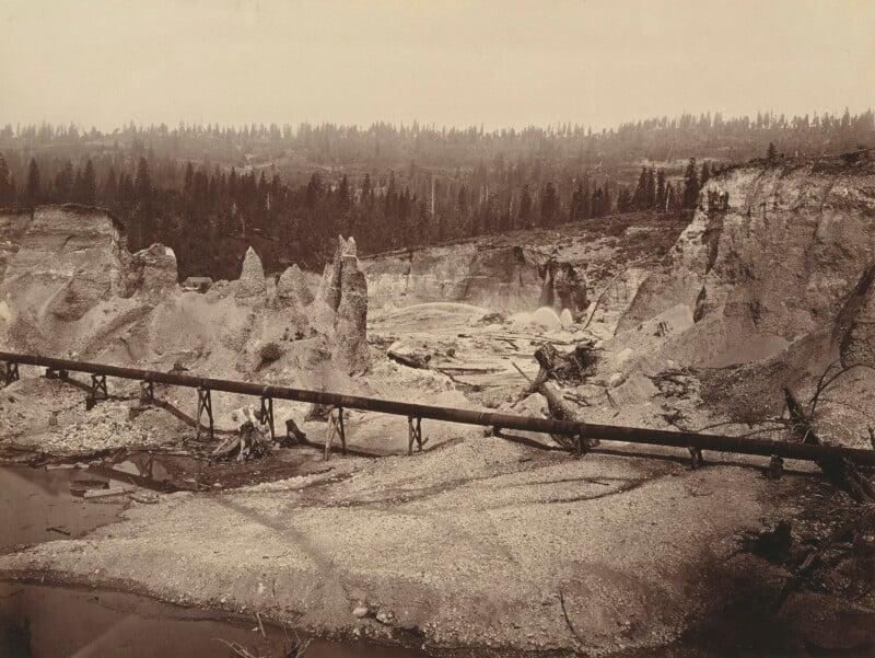 A sepia-toned photo shows a barren, eroded landscape with exposed rock formations and a long wooden pipeline stretching across the scene, surrounded by sparse trees and forested hills in the background.