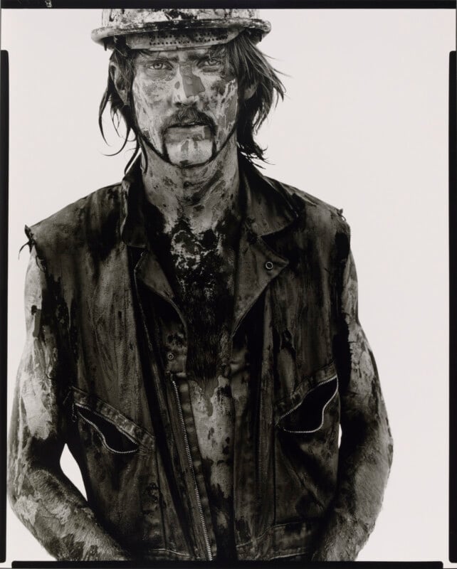 A man wearing a hard hat and a sleeveless vest stands covered in dirt and oil, looking directly at the camera with a serious expression. The image is in black and white.