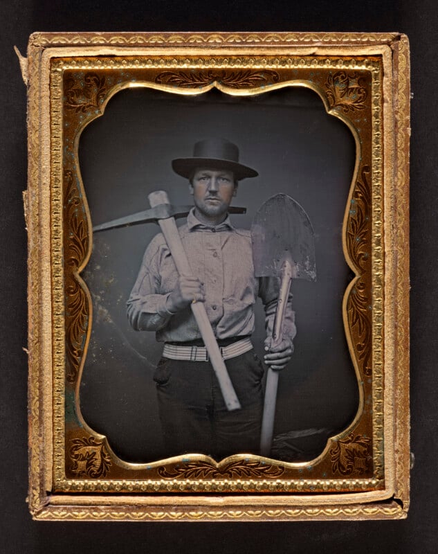 A vintage photograph of a man wearing a wide-brimmed hat and holding a pickaxe and a shovel, framed in an ornate, gold-colored case with decorative patterns.