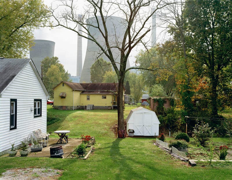 A grassy backyard with a white shed, a picnic table, and lawn chairs. In the background are houses, trees, and large industrial cooling towers partially obscured by mist or fog.