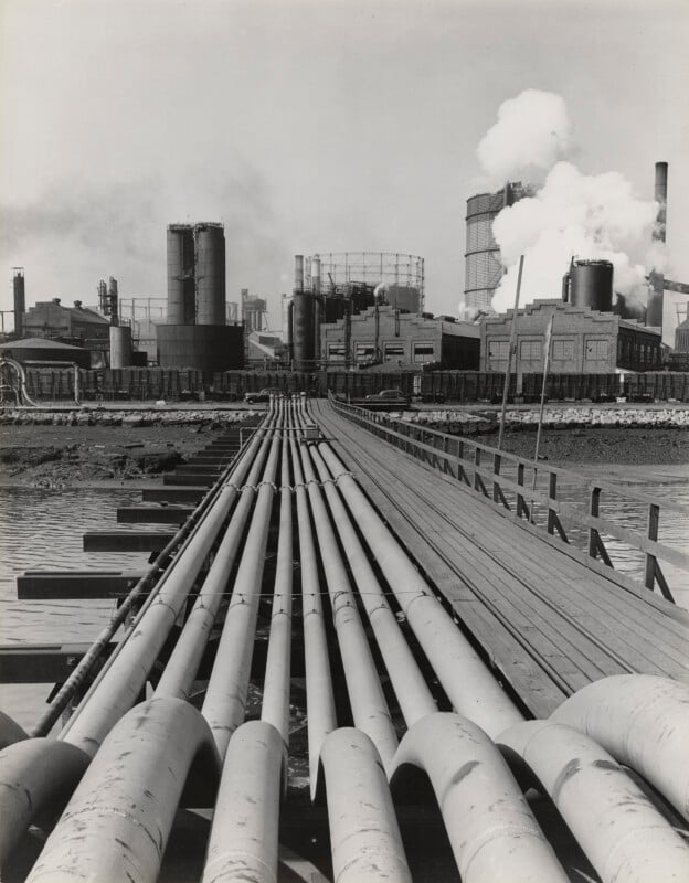 A black-and-white photo shows large industrial pipes leading over water toward a factory complex with smokestacks, cylindrical storage tanks, and steam or smoke rising in the background.