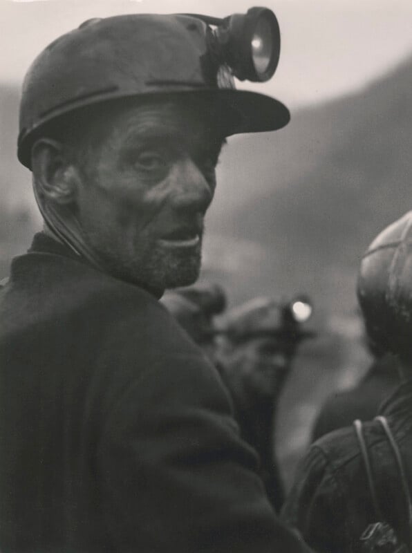 A black-and-white photo of a miner wearing a helmet with a headlamp. His face is dirty, and he looks over his shoulder. Other miners in similar gear are seen in the blurry background.