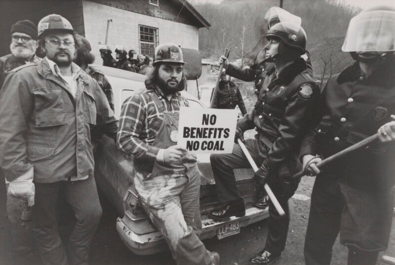 A group of miners and police stand together; one miner in overalls and a hard hat holds a sign reading "NO BENEFITS NO COAL." Police in helmets and uniforms hold batons. A car and building are in the background.