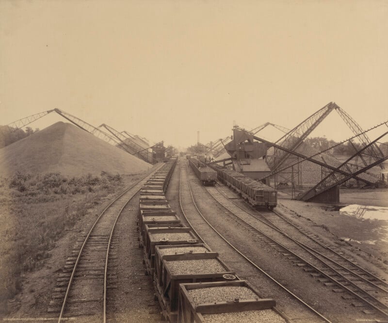 A sepia-toned photo shows multiple railway tracks with trains carrying gravel or coal, large piles of material, and several industrial cranes positioned over the tracks. Trees and a river are visible in the background.