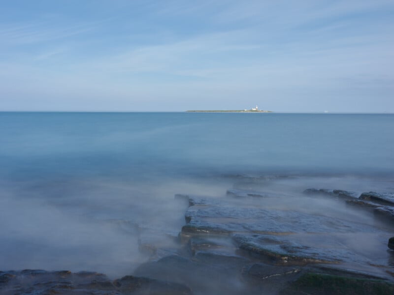 Rocky shoreline with misty water in the foreground, calm blue sea stretching out to a distant small island with a white lighthouse under a blue sky with wispy clouds.