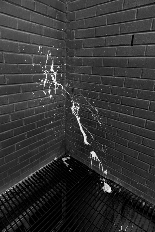 White paint splattered on the corner of two intersecting brick walls above a grated metal floor, shown in black and white. The paint streaks diagonally across the wall, creating an abstract pattern.