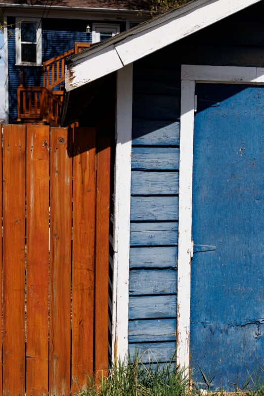 A weathered blue wooden shed with a white trim stands beside a tall, orange-brown wooden fence. Grass grows at the base, and a blue house with a balcony is visible in the background.