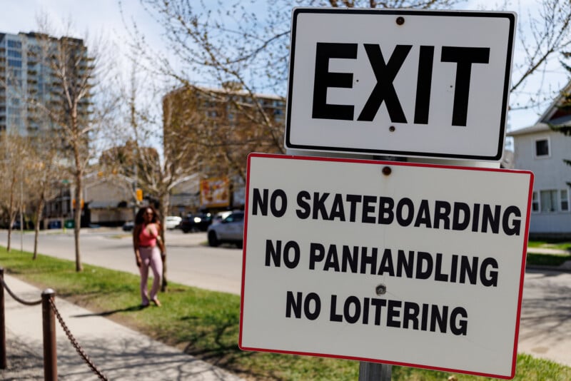 A sign reads “EXIT” above another sign stating “NO SKATEBOARDING, NO PANHANDLING, NO LOITERING.” Trees, buildings, and a person walking on a sidewalk are visible in the background.
