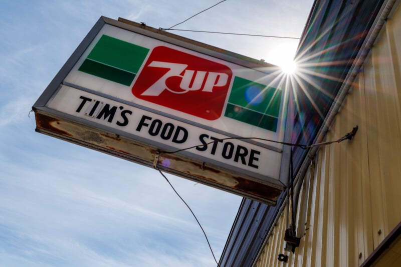 A weathered sign for Tim's Food Store, featuring a 7UP logo, hangs on a building with yellow siding as sunlight creates a starburst effect in the sky.