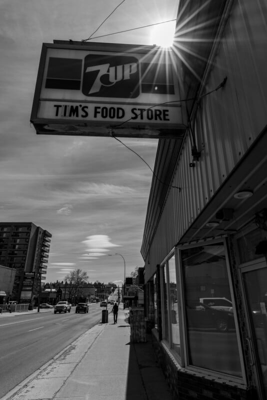 Black and white photo of a street scene with a large 7UP sign reading “Tim’s Food Store” on a building. The sun shines brightly above, and two people walk on the sidewalk in the distance.