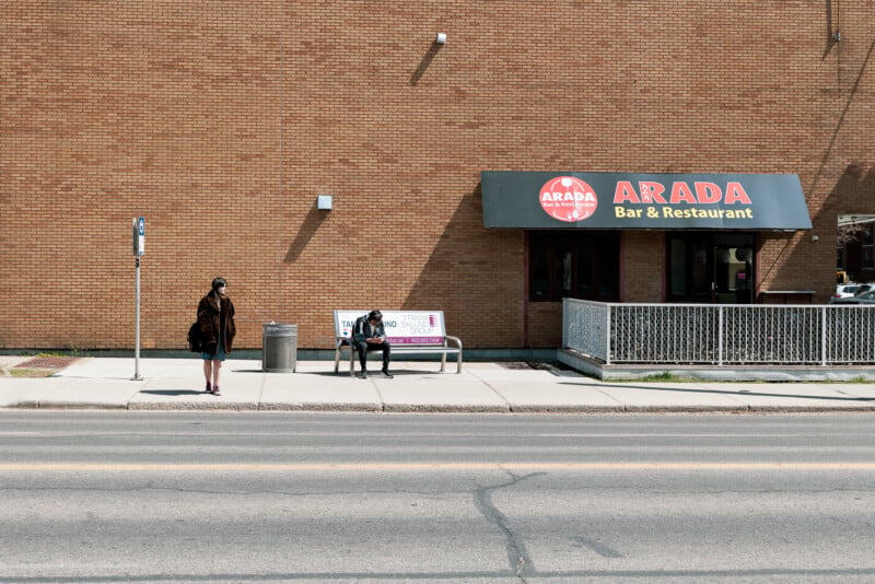 Two people wait at a bus stop in front of a brick building with an "Arada Bar & Restaurant" sign. One person stands near the curb; the other sits on a bench next to a trash can. The street is empty.