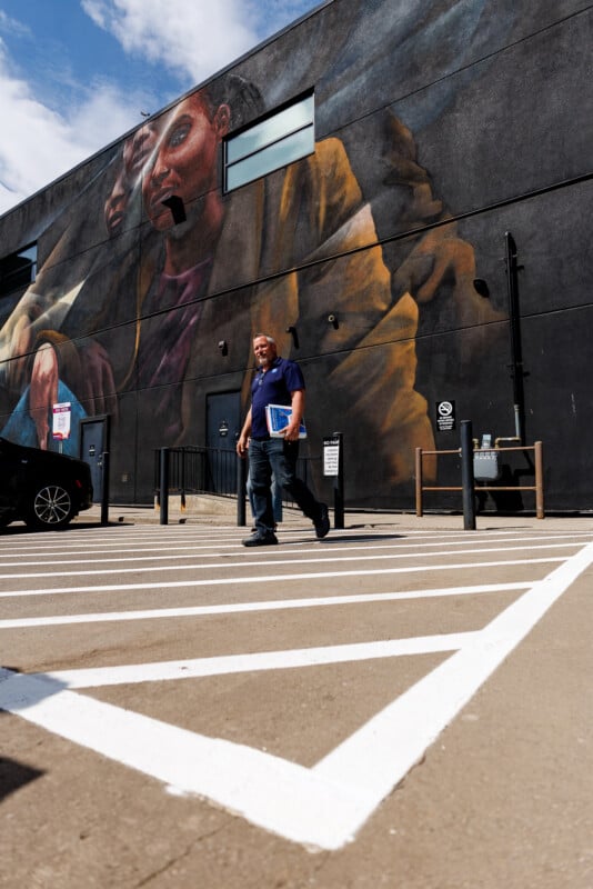 A man carrying folders walks through a parking lot in front of a large, detailed mural painted on a dark building wall under a partly cloudy sky.
