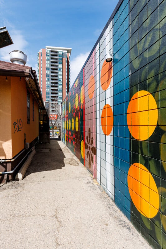 A narrow alley with colorful, geometric mural featuring orange circles on a tiled wall, next to a plain tan building; a tall apartment building is visible in the background under a blue sky.