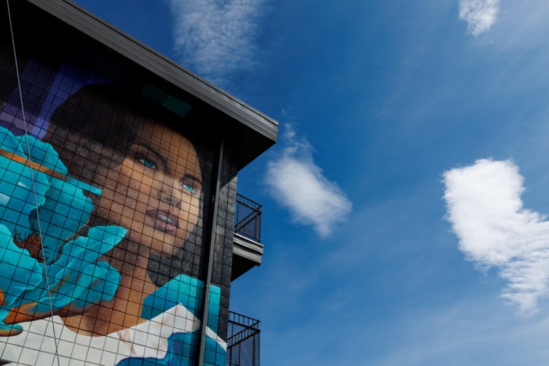 A colorful mural of a woman's face with blue eyes and blue shapes around her is painted on the side of a building, set against a bright blue sky with scattered white clouds.