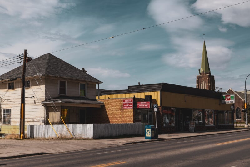 A street view of an old residential house next to a small convenience store with a 7-Eleven sign. A church with a pointed steeple rises in the background under a partly cloudy sky.