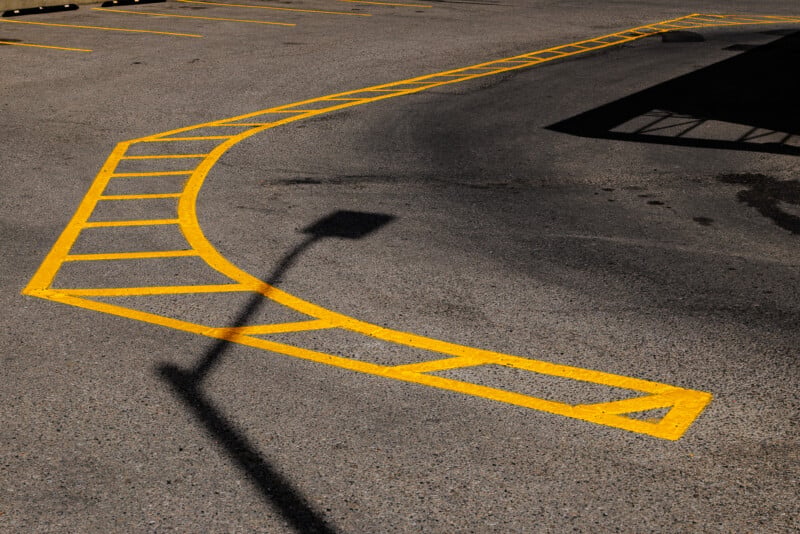 A nearly empty parking lot with a curved, diagonal yellow grid line painted on the asphalt, and the shadow of a streetlamp stretching across the pavement.
