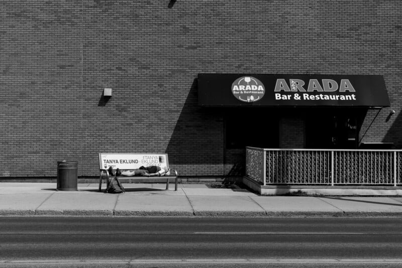 A person lies on a bench next to a trash can outside the Arada Bar & Restaurant, which has a large black awning, against a brick wall on an empty sidewalk.