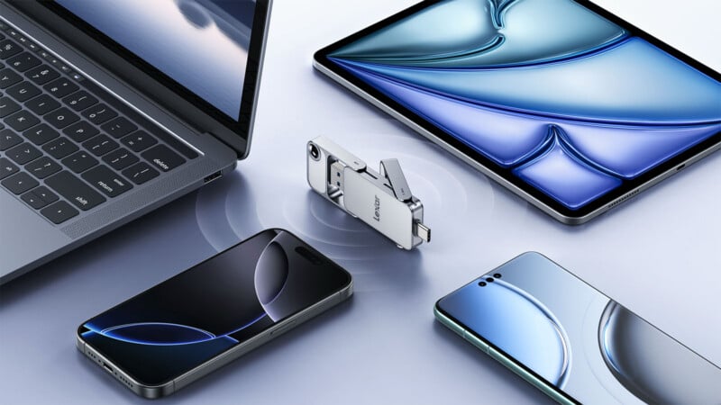 A multi-port flash drive is placed on a table, surrounded by a laptop, a tablet, and two smartphones. The devices display blue abstract patterns on their screens.