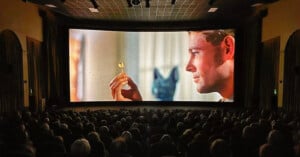 A crowded movie theater audience watches a large screen showing a close-up of a man holding a gold object, with an ancient Egyptian statue in the background.