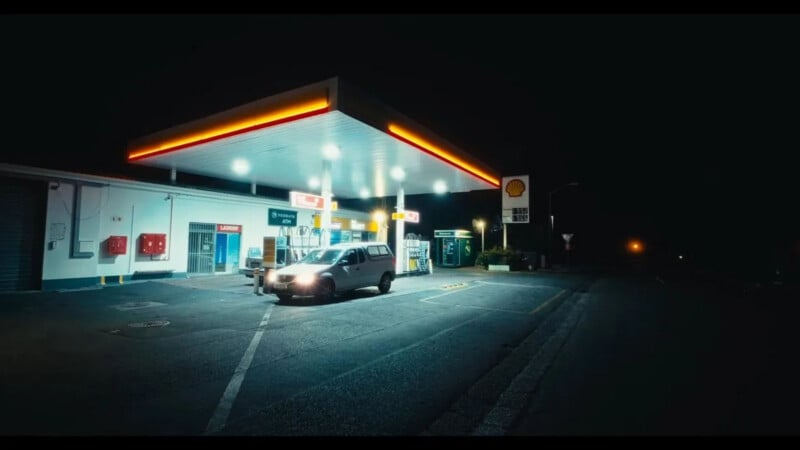 A car is parked at a brightly lit Shell gas station at night, surrounded by darkness. The area is mostly empty, with vivid lights illuminating the pumps and canopy.