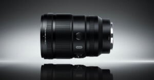A black camera lens is positioned horizontally on a reflective surface against a gradient gray background, displaying its details and metallic lens mount.