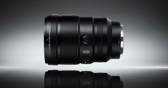 A black camera lens is positioned horizontally on a reflective surface against a gradient gray background, displaying its details and metallic lens mount.