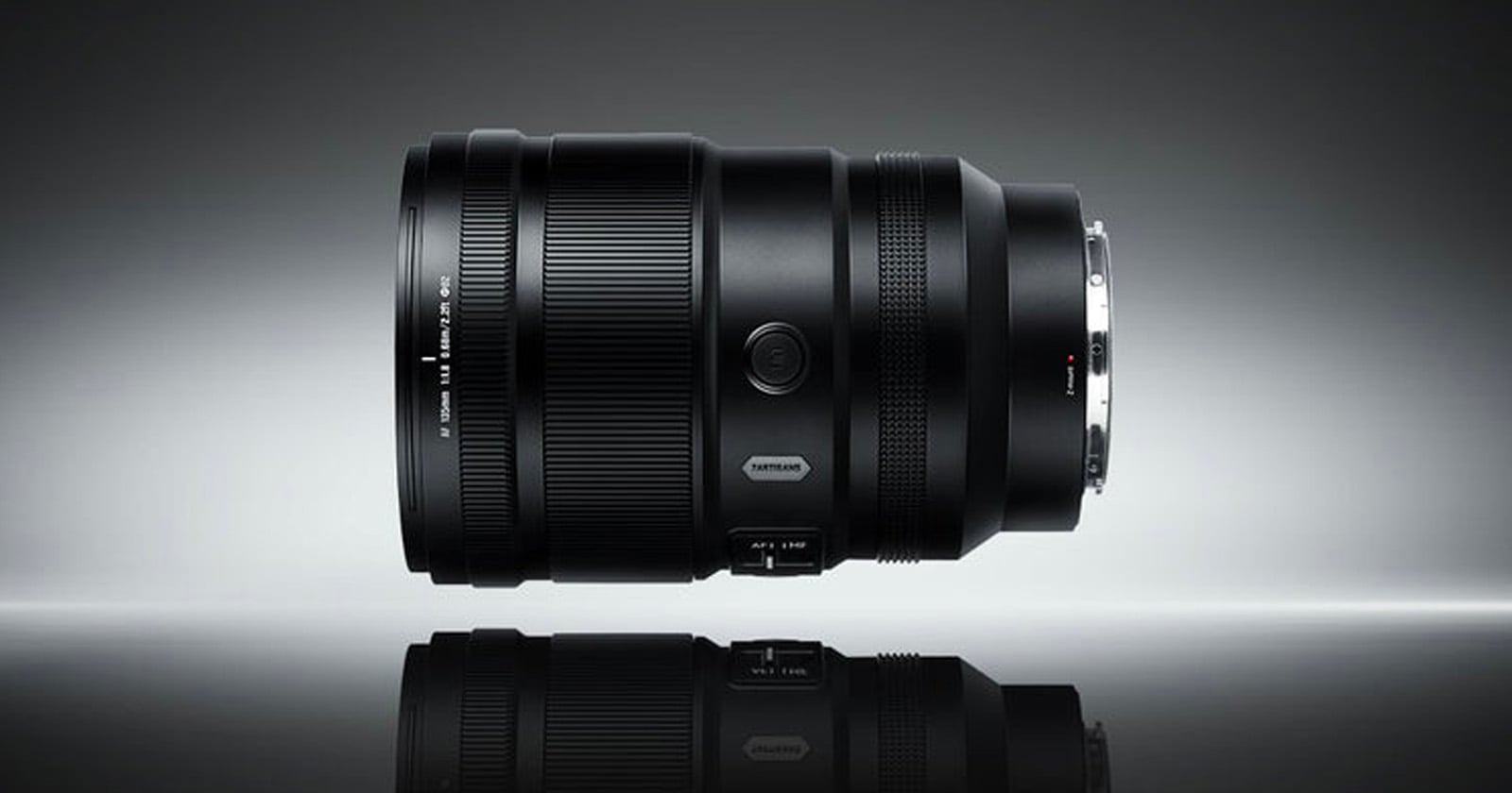 A black camera lens is positioned horizontally on a reflective surface against a gradient gray background, displaying its details and metallic lens mount.
