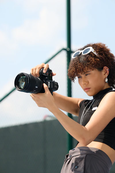 A young woman with curly hair and sunglasses on her head is outdoors, focusing intently while holding and adjusting a professional camera. A blurred fence and blue sky are in the background.