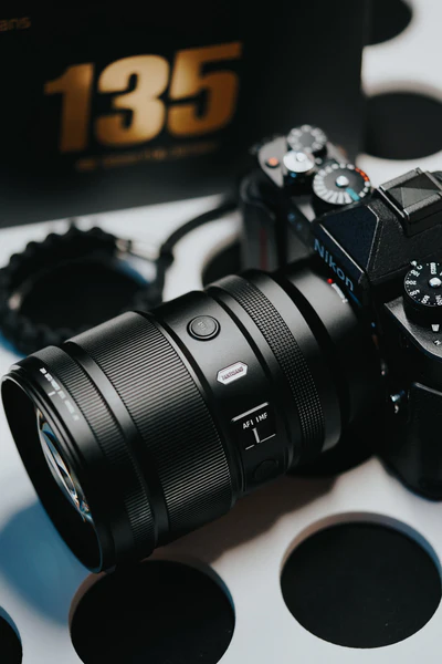 Close-up of a Nikon camera with a large lens resting on a white surface with black circles, and a blurred box featuring the number “135” in bold gold letters in the background.