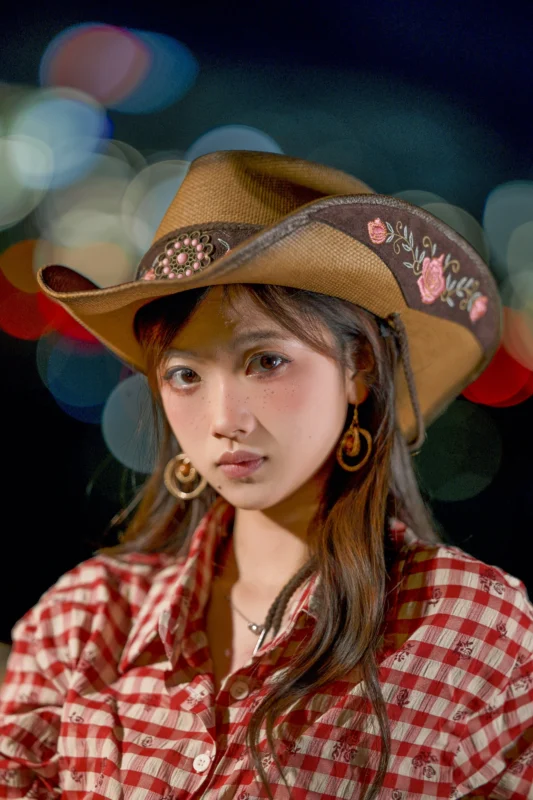 A young woman wearing a brown cowboy hat with floral embroidery and a red checkered shirt stands against a blurred, colorful night background. She has long brown hair and wears hoop earrings.