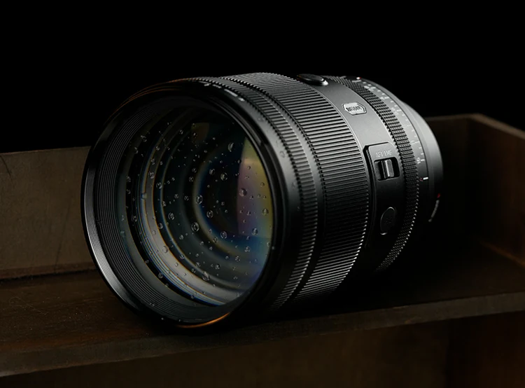A close-up of a camera lens with water droplets on the glass, resting on a wooden surface against a dark background.