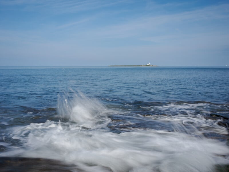 Waves crash onto rocks in the foreground, with calm blue sea stretching to the horizon and a small island with a lighthouse visible in the distance under a blue sky.