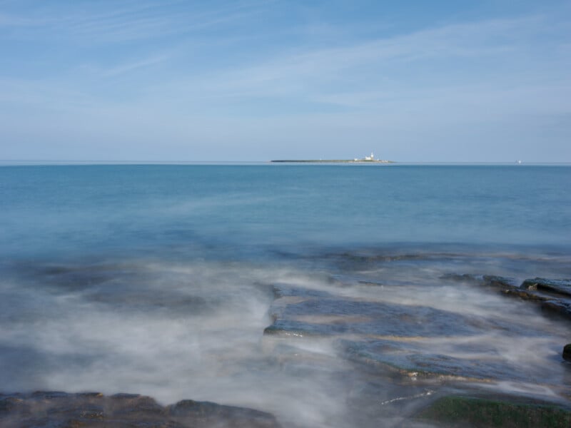 Rocky shoreline with gentle waves in the foreground, a calm blue sea, and a distant low-lying island with a lighthouse under a clear, light blue sky.
