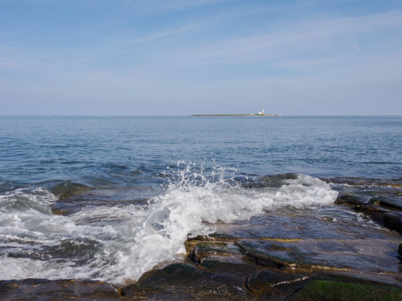 Waves splash against rocky shore under a blue sky, with a small distant island featuring a white lighthouse visible on the horizon.