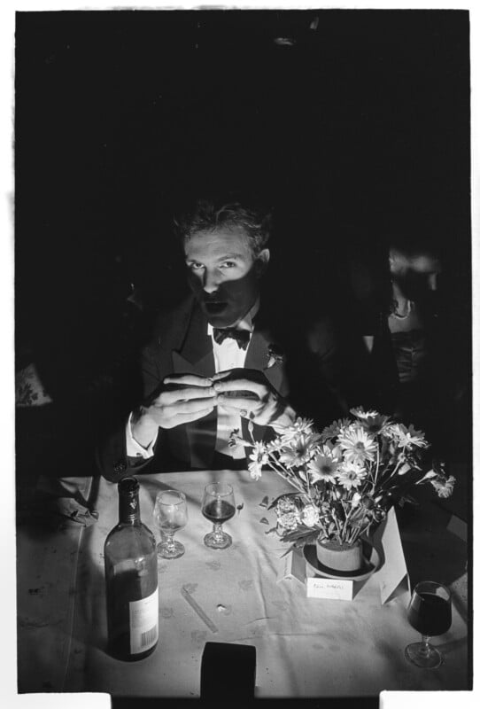 A man in a suit and bow tie sits at a table with a bottle of wine, glasses, and a bouquet of flowers, dramatically lit with shadows obscuring the background. He appears to be holding a piece of bread or food.