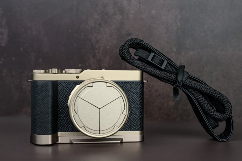 A modern digital camera with a textured black and silver body sits on a surface, featuring a geometric lens cover; a black camera strap is coiled neatly beside it.
