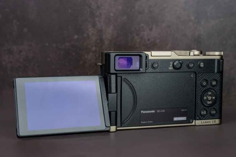 A digital camera with a flip-out LCD screen is shown from the back. The screen is extended to the left, and various control buttons and a viewfinder are visible on the right side. The camera is on a brown surface.