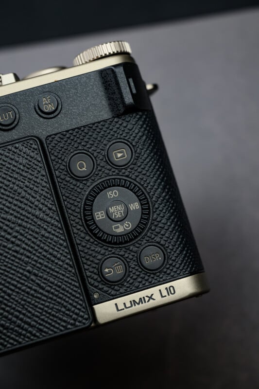 Close-up of the control buttons and textured grip on a black and gold Panasonic Lumix L10 camera, showing the menu dial, display button, and part of the top dials.
