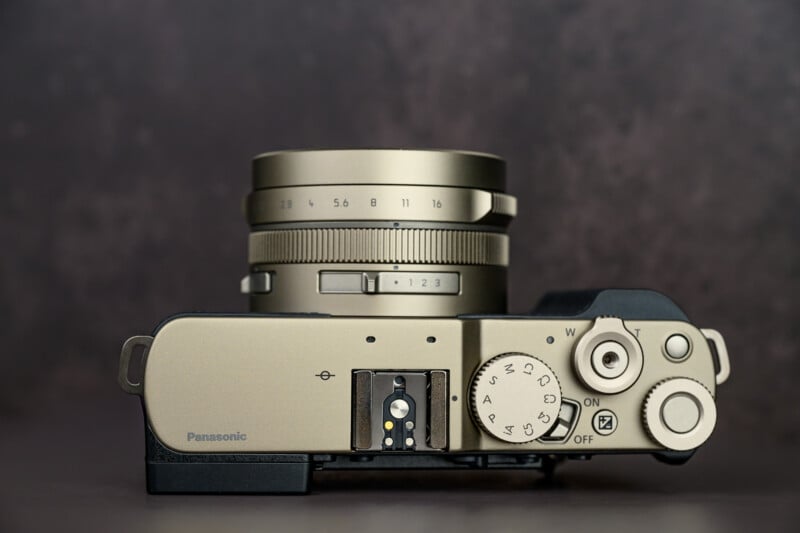 Top view of a Panasonic digital camera in a metallic finish, showing lens, dials, buttons, and the on/off switch against a blurred dark background.