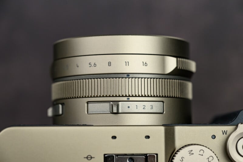 Close-up of a metallic camera lens with visible aperture settings (from f/2 to f/16), textured focus ring, and a switch labeled 1, 2, 3 on a beige camera body.
