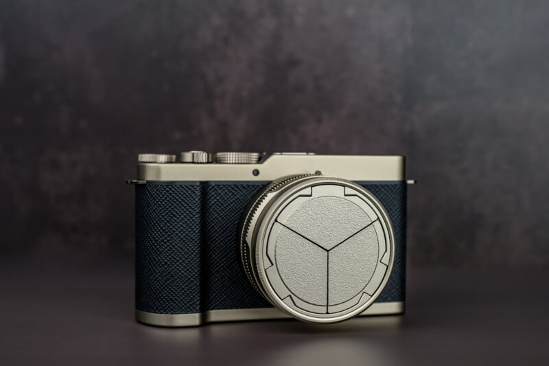 A stylish, modern digital camera with a textured black body and metallic silver accents sits on a dark surface against a blurred, neutral background. The lens cap features a unique geometric pattern.